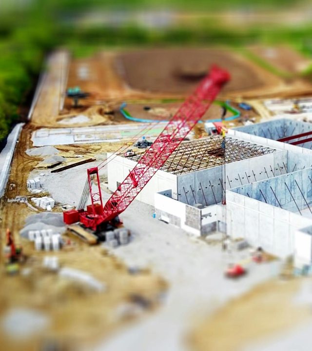 Aerial view of a construction site with a red crane amidst green fields and concrete buildings.